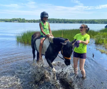 Camper wearing a helmet riding a horse in the water with staff guiding the horse for safety